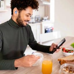 Young man with a hearing aid looks at his smartphone while having breakfast at home.