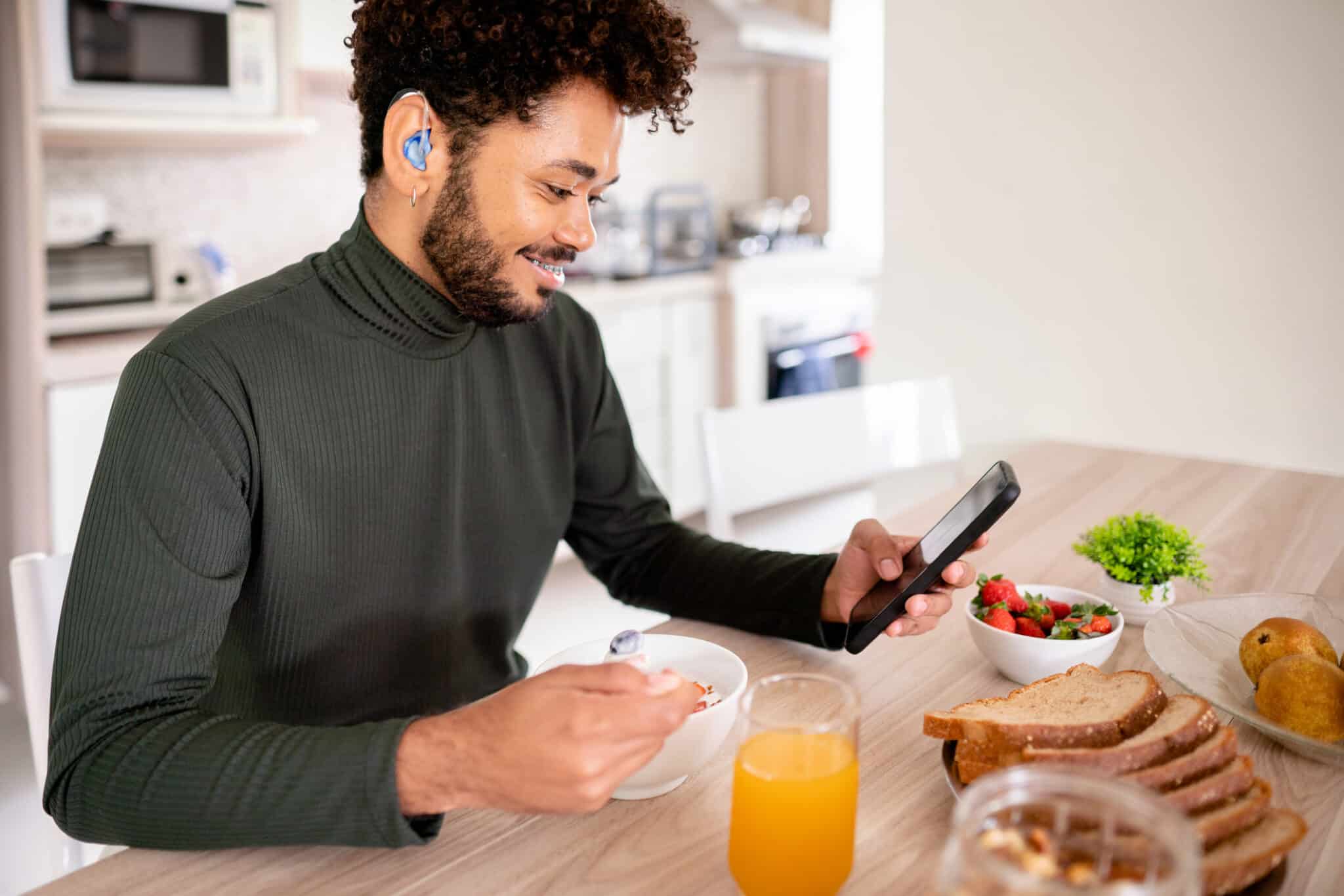 Young man with a hearing aid looks at his smartphone while having breakfast at home.