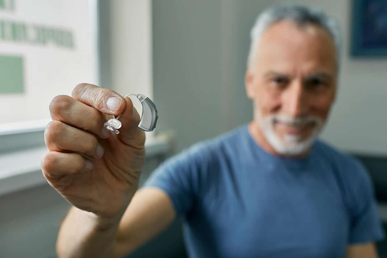 Senior man holding BTE hearing aid in hand on foreground, close-up.