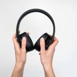 A pair of over-ear headphones held up by a pair of hands in front of a pale background.